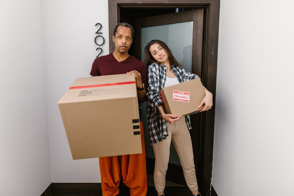 A man and a woman stand inside a building near an open door, each holding cardboard boxes used for home relocation. The man, wearing an orange pair of trousers and a maroon t-shirt, holds a large card