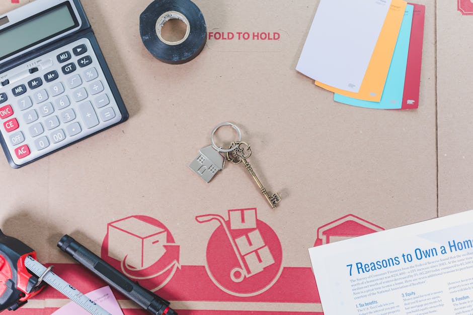 A close-up view of a packaging and moving workspace with various tools and materials arranged on a flat surface. Visible are a calculator, a roll of black tape, a set of colour-coded file folders, a m