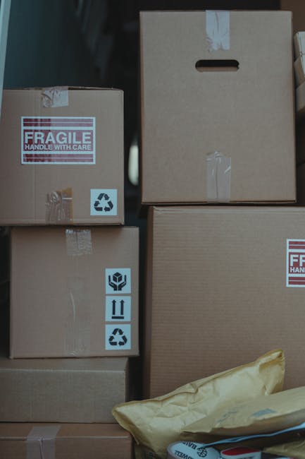 A collection of cardboard boxes of various sizes stacked inside a property, prepared for a home relocation or goods transport. Some boxes feature handling labels such as 'Fragile' and recycling symbol